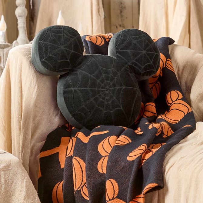 Black Mickey Mouse-shaped pillow with spiderweb pattern rests on an orange-and-black blanket featuring pumpkin designs. Neutral fabric backdrop.