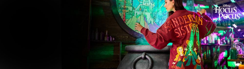 A person wearing a burgundy sweatshirt featuring a design of the Sanderson Sisters from Hocus Pocus near a cauldron with a vibrant backdrop.