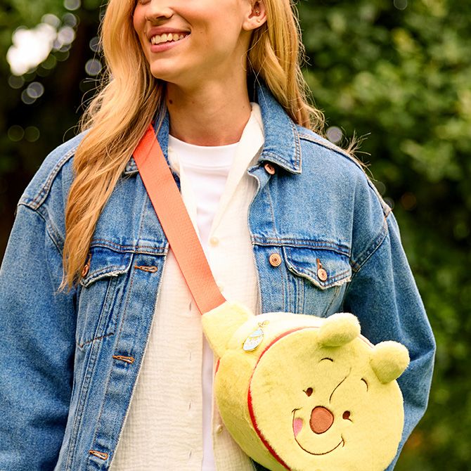 Smiling woman in a denim jacket carries a Winnie the Pooh plush crossbody bag with an orange strap. Trees and brick building in the background.
