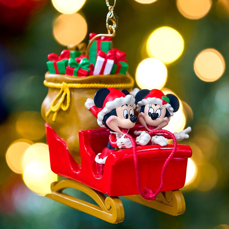 Mickey Mouse and Minnie Mouse, dressed in festive Santa hats, ride a red sleigh with a sack of gifts against a blurred holiday light background.