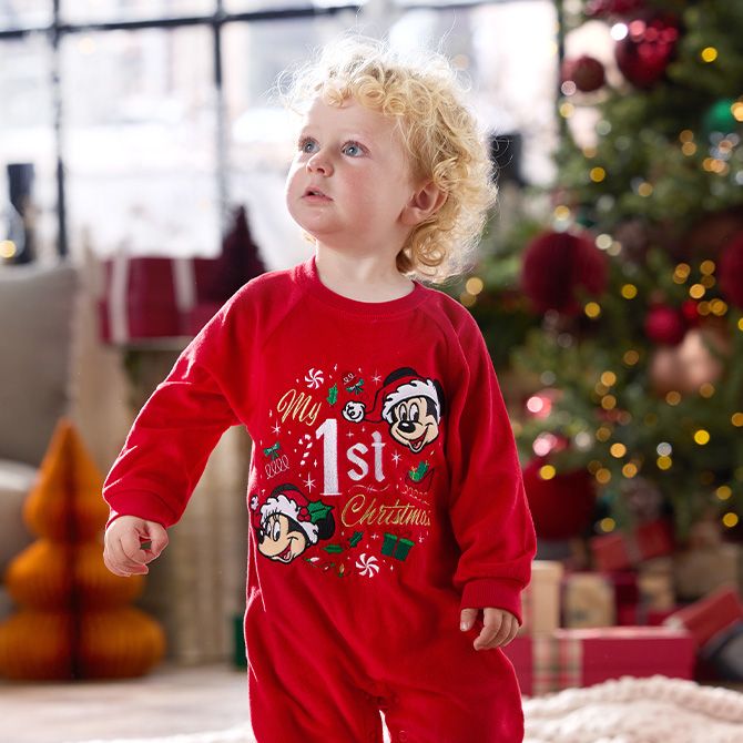 Toddler wearing a red onesie with 'My 1st Christmas' text and Mickey Mouse graphics, standing near a decorated Christmas tree and gifts.