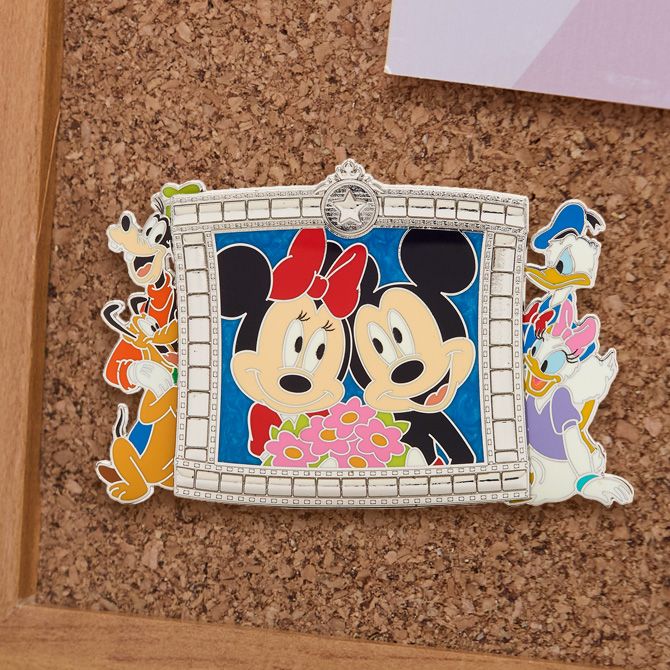 Enamel pin of Mickey and Minnie Mouse smiling in a jeweled silver frame, with Goofy, Donald, Daisy, and Pluto around the edges, pinned on a cork board.