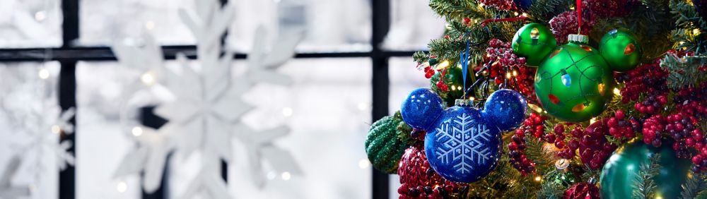 Green and blue Mickey Mouse Christmas ornaments with a snowflake and holiday lights design hang on a decorated tree with berries and white lights.