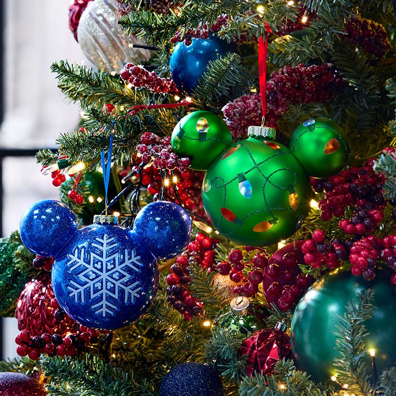 Green and blue Mickey Mouse Christmas ornaments with a snowflake and holiday lights design hang on a decorated tree with berries and white lights.