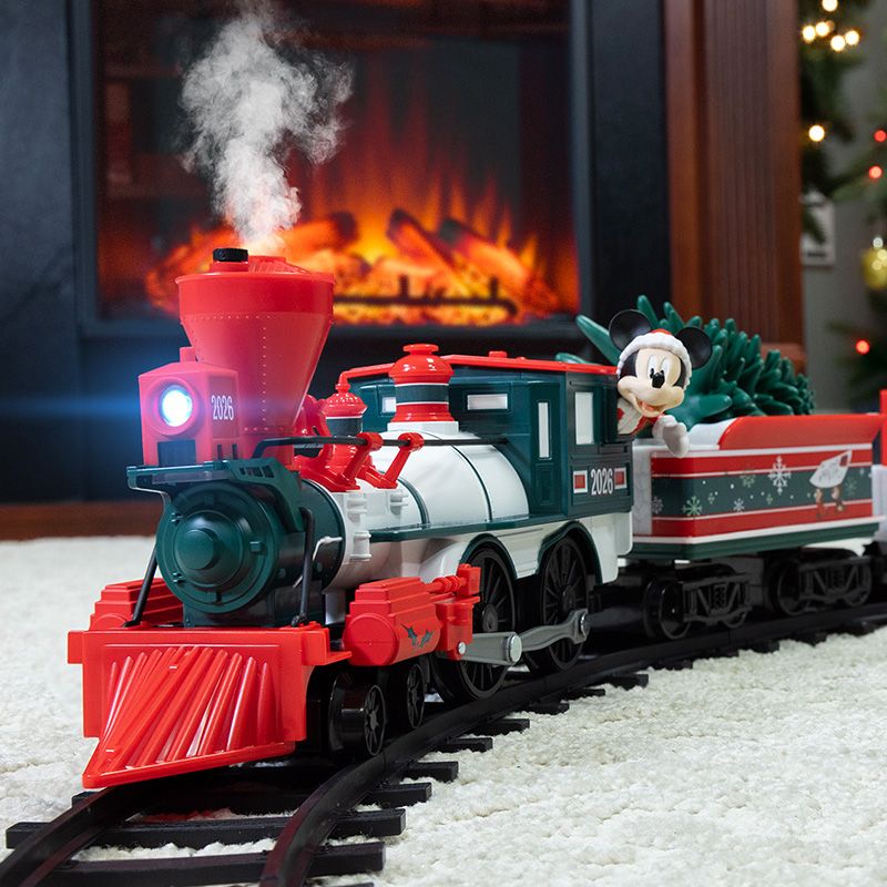 Toy train with "2026" on the engine, emits steam as it circles a track. Minnie Mouse rides in a festive train car with a Christmas tree. Fireplace in background.