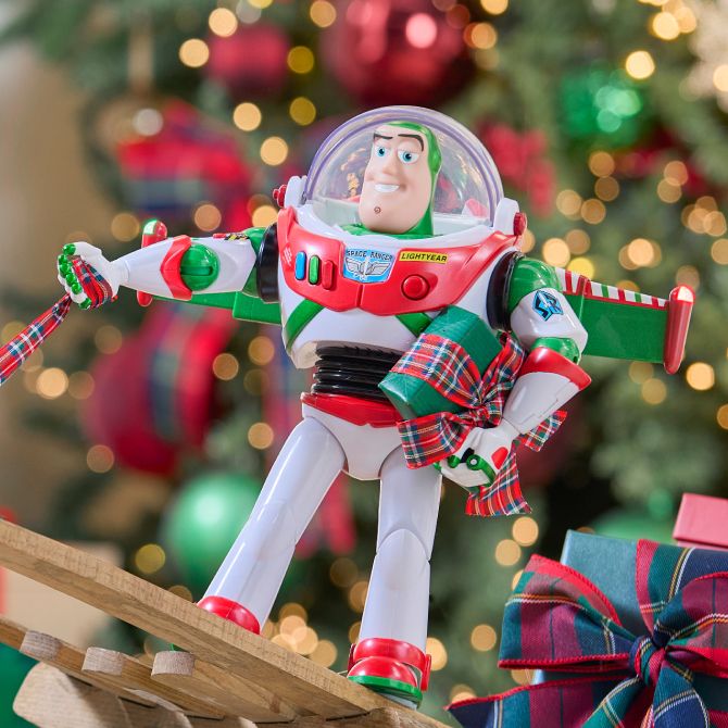 Buzz Lightyear action figure with a plaid ribbon stands before a decorated Christmas tree with ornaments and wrapped gifts in the background.