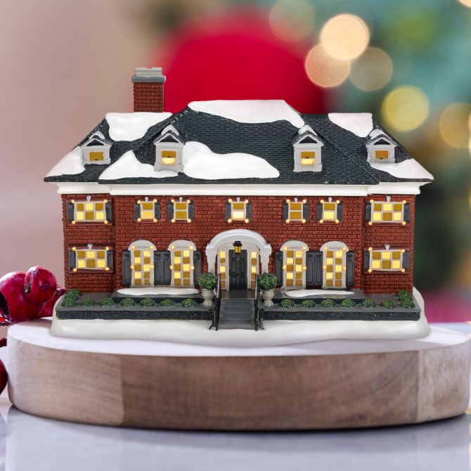 Snow-covered red brick house ornament with a red ribbon is displayed on a wooden base, with blurred holiday lights in the background.