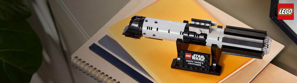 LEGO Star Wars Darth Vader&rsquo;s lightsaber model on a black display stand sits atop stacked notebooks with the LEGO logo in the top right; desk background.
