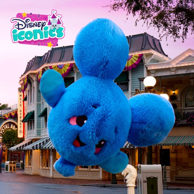 Large blue plush in the shape of Mickey Mouse&rsquo;s head floats above Main Street with the Disney Iconics logo. Pink sky and decorated buildings in background.