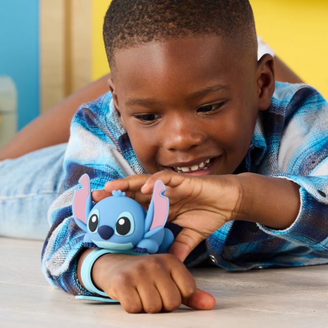 Smiling boy in a blue flannel shirt plays on the floor with a blue Stitch toy. The background is colorful with yellow and blue walls.