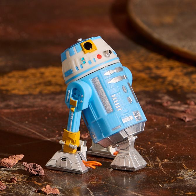 Blue and white Star Wars droid figure with yellow details stands on a rusty, textured metal surface with scattered debris in the background.