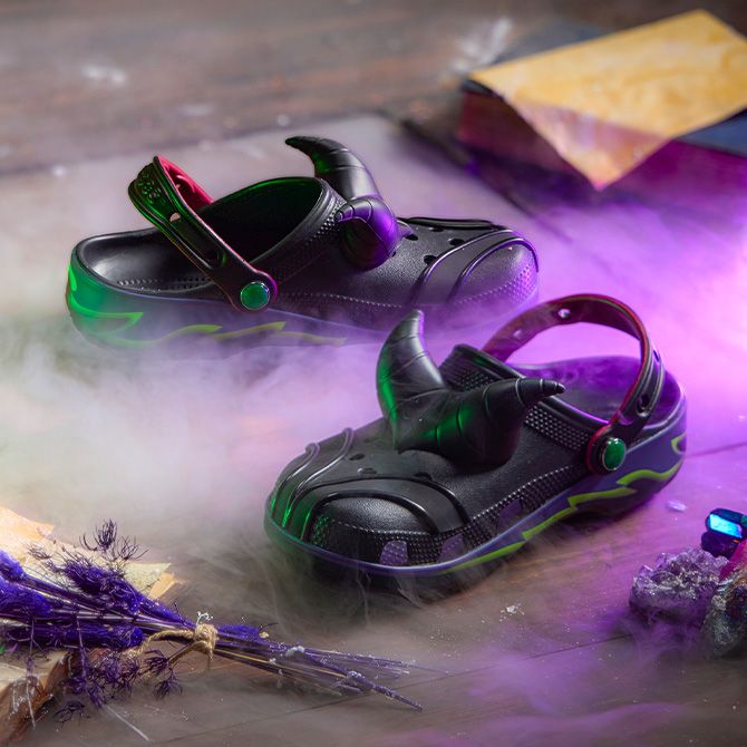 Black slip-on shoes inspired by Maleficent with horn details, glowing green and purple highlights, placed on a misty wooden surface with spellbook and dried flowers.