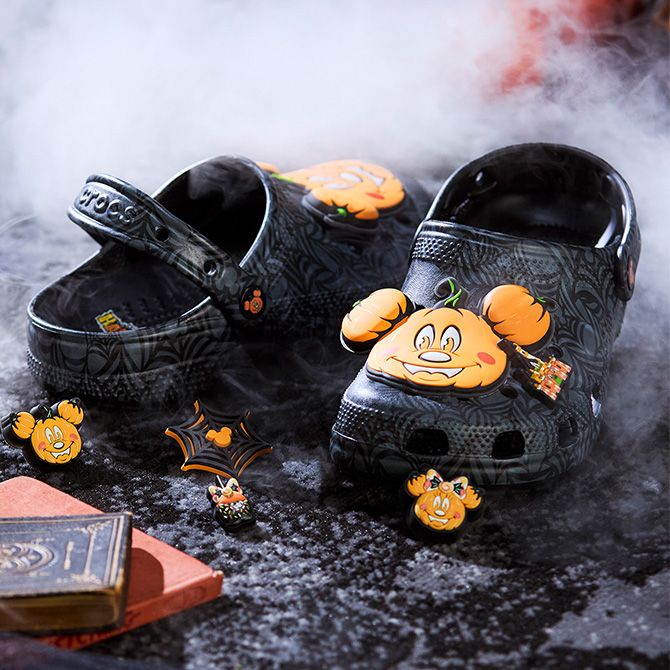 Black Crocs with Mickey pumpkin charms and bat decorations are set on a misty, textured surface, with a book in the foreground and a smoky background.