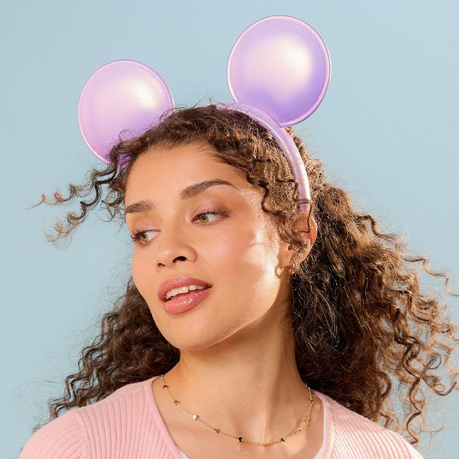 Woman wears shiny lavender Mickey ear headband, soft pink top, and gold necklace against a plain light blue background.