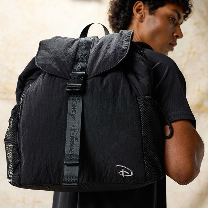Person carries a large black Disney backpack featuring mesh side pockets and a Disney logo patch, with a neutral marble-tone wall behind.