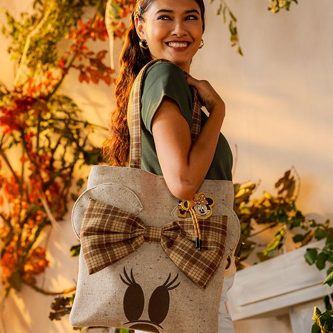 Woman smiles while holding a beige Daisy Duck tote bag with a large plaid bow and Minnie Mouse keychain. Fall leaves and warm light are in the background.