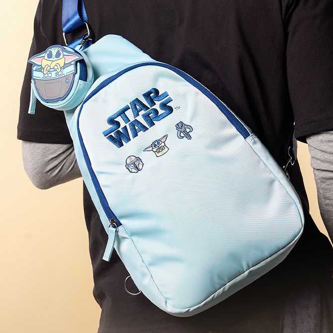 Person wears a light blue Star Wars sling bag with Grogu, Mandaorian helmet, and droid graphics and a Grogu round coin pouch; beige background.