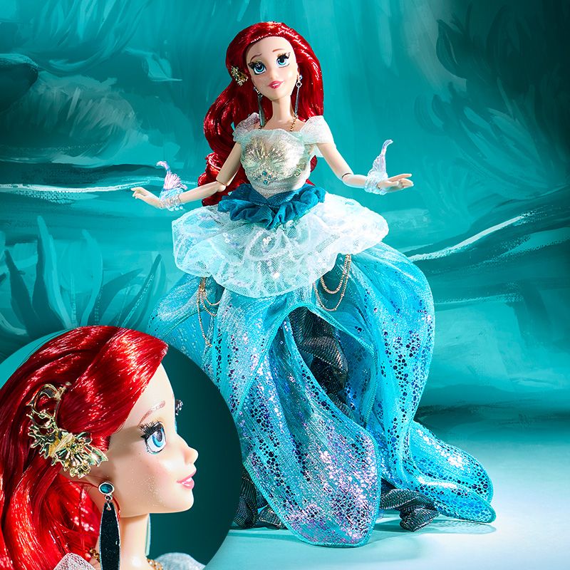 Jasmine doll wears a shimmering blue gown with gold accents and holds a white flower, posed in the snow with evergreens and soft holiday lights.