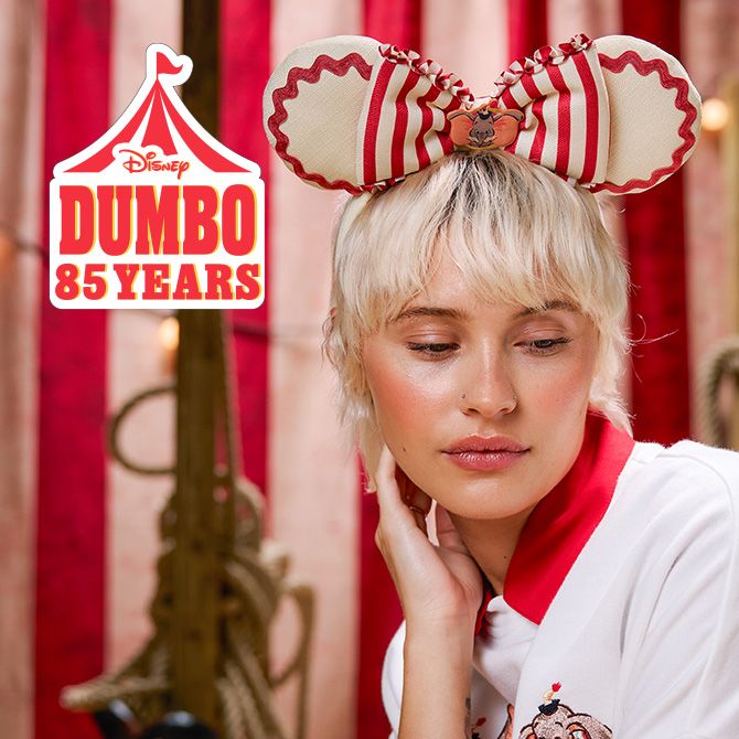 A woman wearing Dumbo mouse ears and a white polo with a red collar sits before red and white circus stripes with glowing string lights.