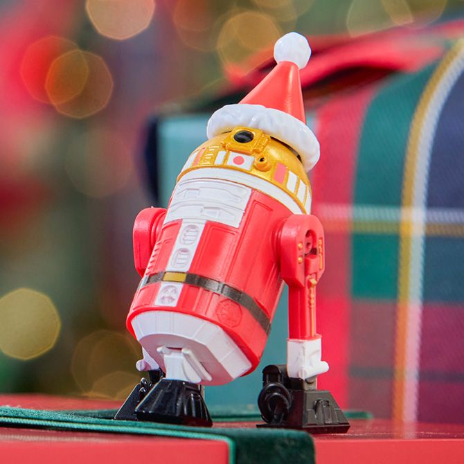 Red and white Star Wars droid figure dressed as Santa with a Santa hat stands in front of plaid-wrapped gifts and holiday lights.