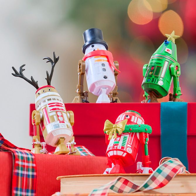 Four Star Wars droid figures dressed as a reindeer, snowman, Christmas tree, and holiday gifts are displayed on wrapped presents with festive lights.