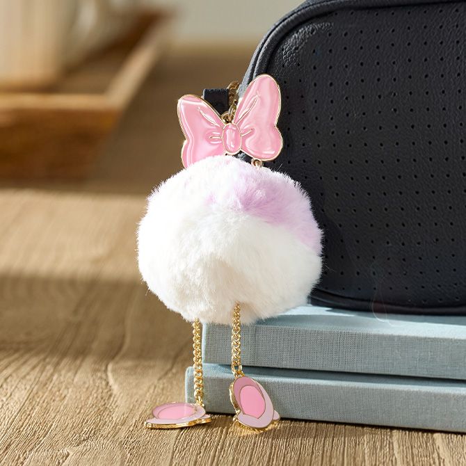 Fluffy white keychain with a pink bow, gold chain legs, and pink shoes hangs on a black bag with wood surface and soft background.