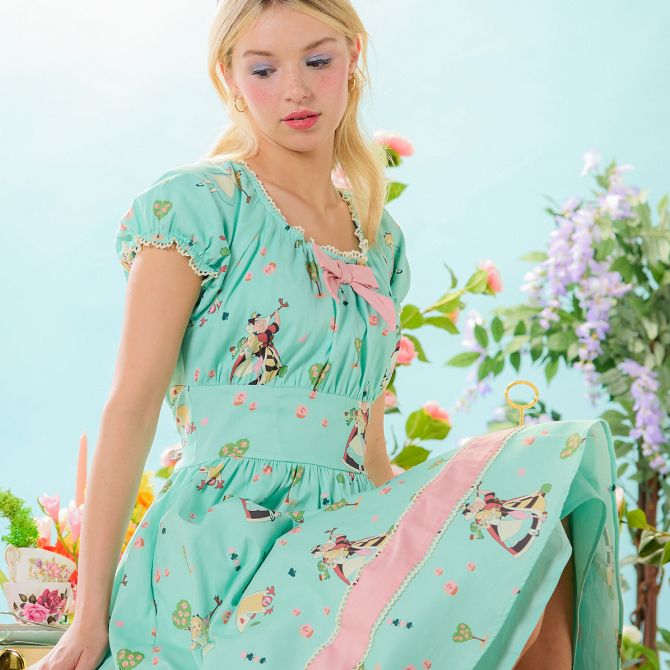 Woman in a mint green dress with Alice in Wonderland print and pink bow sits in a pastel garden setting with flowers.