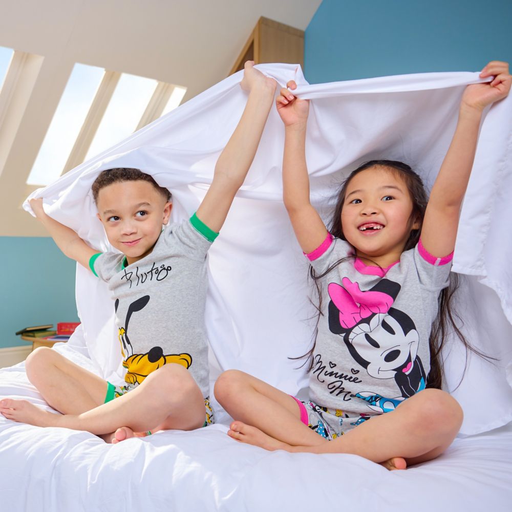 Two children wearing festive Mickey Mouse and Disney-themed pajamas sit near a decorated Christmas tree holding plush toys.