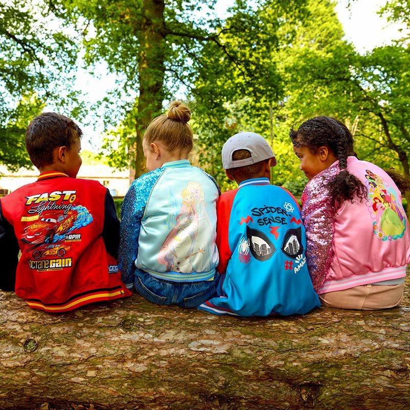 Four children sit on a log wearing colorful jackets featuring Cars, Frozen, Spider-Man, and Disney Princess designs outdoors in a park setting.