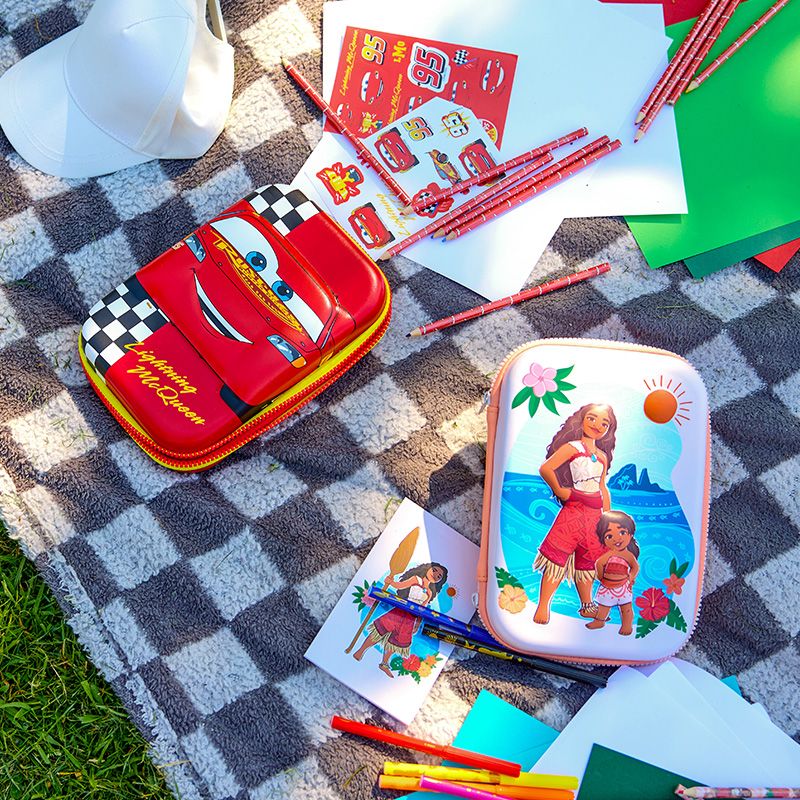 On a checkered blanket, colorful Disney-themed stationery includes a Lightning McQueen pencil case and Moana-themed art supplies surrounded by paper.