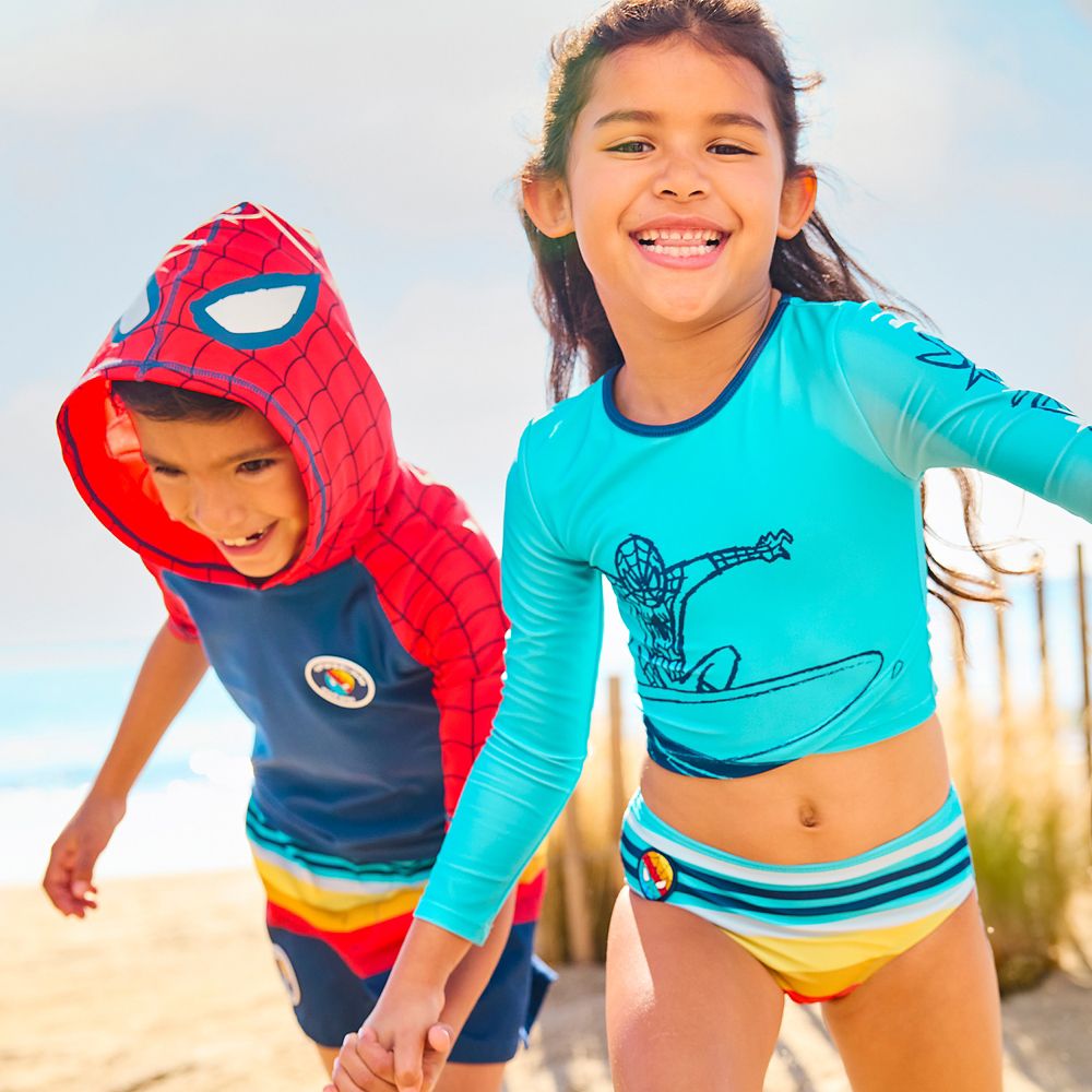 Young boy wearing a hooded Spiderman swim shirt and trunks and young girl wearing a teal Spiderman swimsuit on a beach.