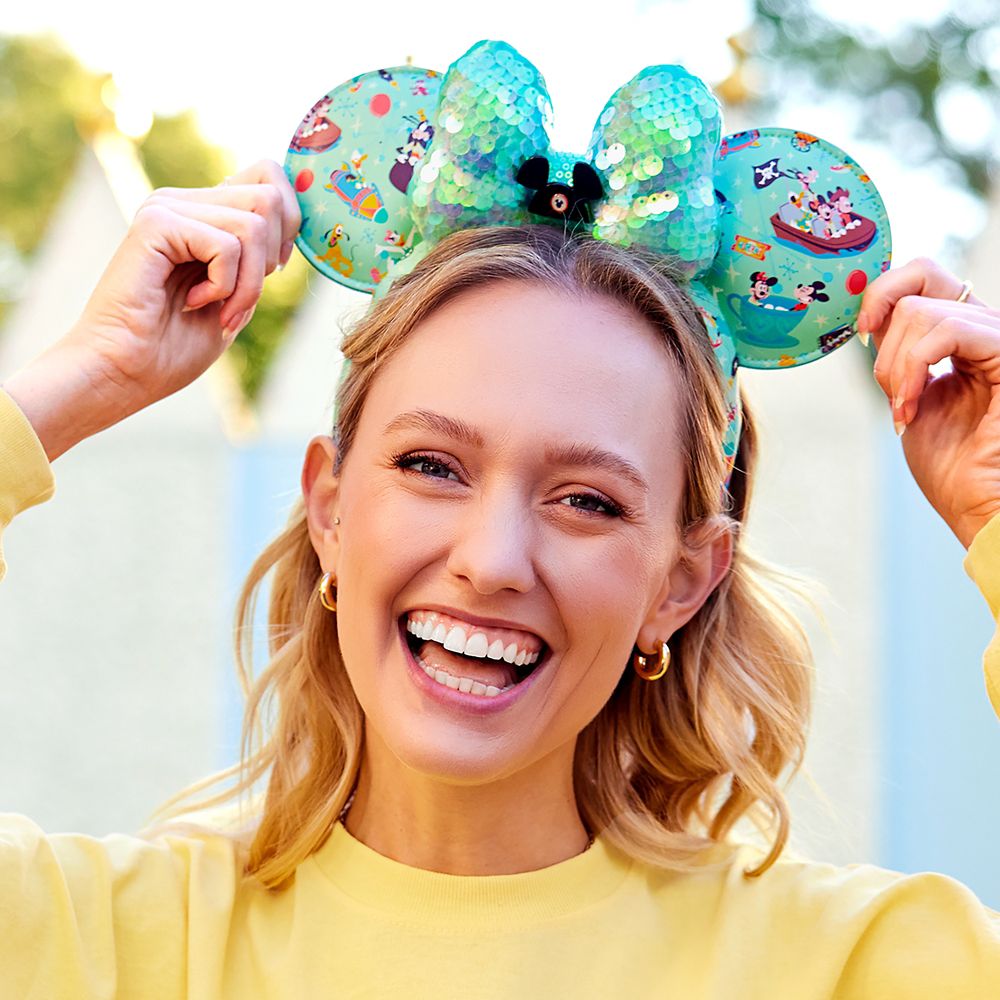 Blonde woman wearing a yellow long sleeve shirt and teal "Play in the Park" mickey ears.