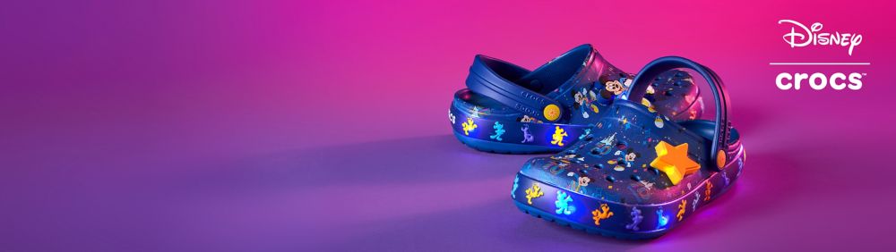 Disney x Crocs blue shoes featuring Mickey Mouse and friends with colorful designs and star-shaped Jibbitz charms on a pink background.